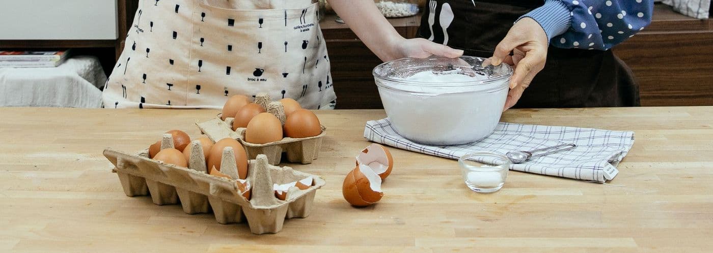 Crop unrecognizable females wearing aprons touching bowl with fresh whipped cream placed on table near eggs in modern kitchen